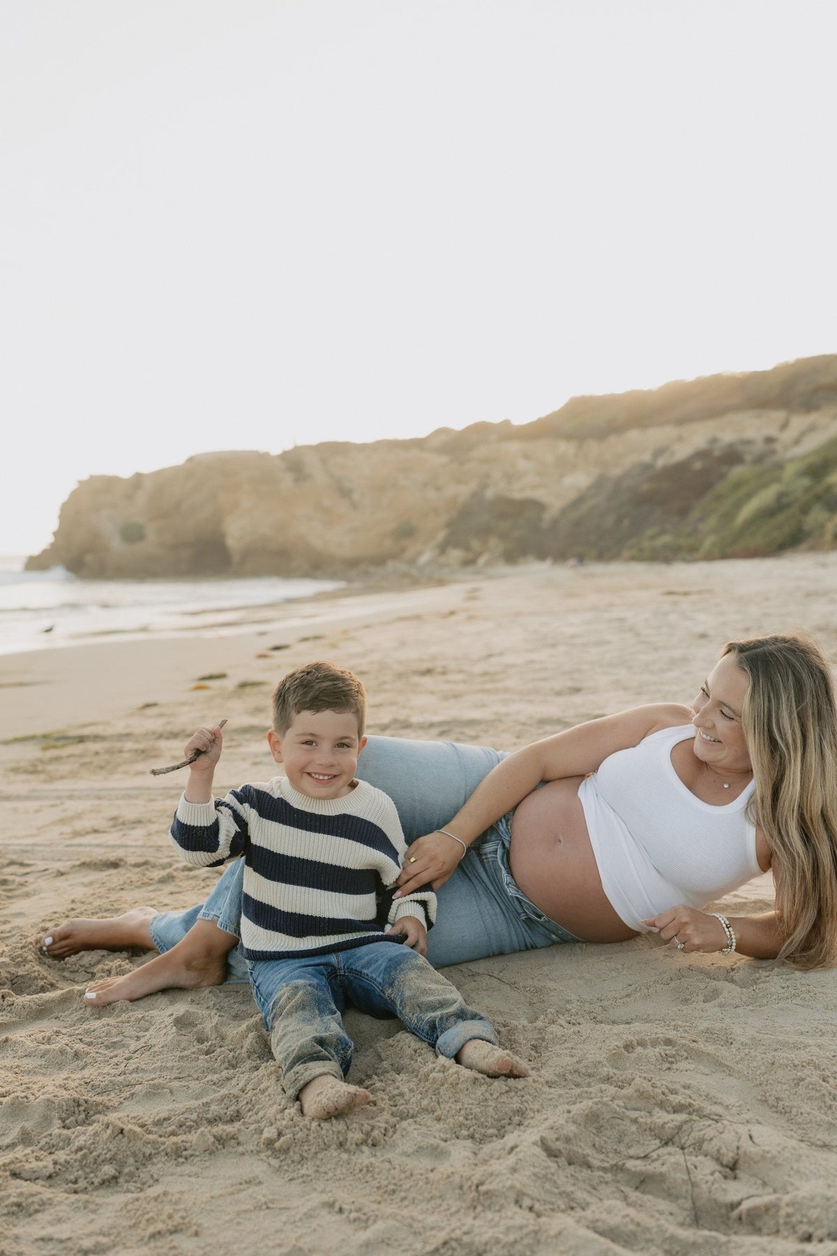 Bridger grinning at the camera while his pregnant mom Makenzie lies on the sand beside him